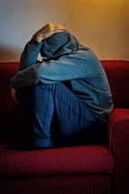 Young man is sitting alone on a couch, curled up with his head in his hands, suffering from depression