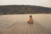 a boy child in a raincoat sitting on a dock 