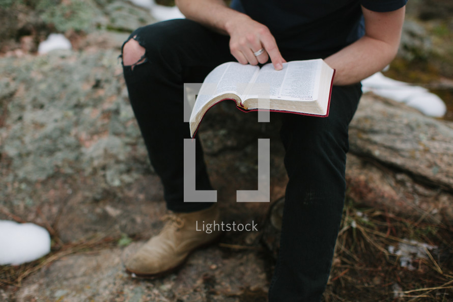 a man sitting on a rock reading a Bible outdoors 