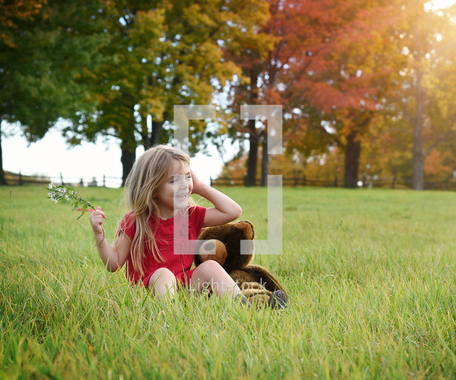 a happy little girl sitting in grass with her teddy bear 