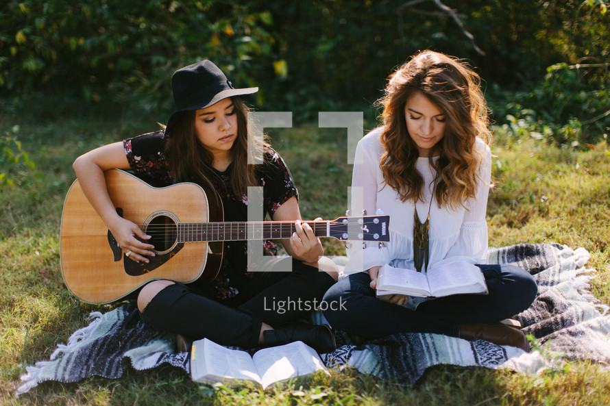 teen girls sitting on a blanket outdoors playing a guitar and singing 
