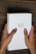 A man's hands holding a white Bible on a wooden table.