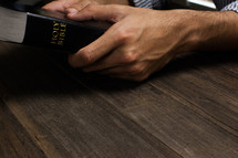 Hands holding onto a Bible on a wooden table.