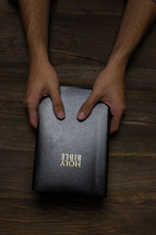 Two hands holding a Bible on a wooden table.