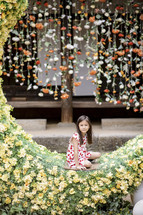A young girl sitting in a crescent moon with a flower wall behind her
