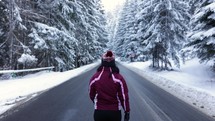 Girl Wearing Winter Clothes Walking On Isolated Road Passing By Dense Forest Covered With Snow. Dolly Shot
