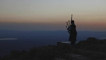 Man playing the bagpipes standing on a cliff watching the sunset