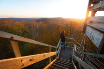 Adventurous young man soaks in the sunrise on stairs of wooden watchtower over autumn foliage trees in Bickle Knob West Virginia
