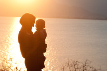 mother holding a toddler in front of a lake under a sunset