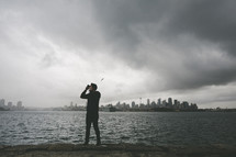 man standing across the river from a city taking a picture 