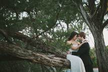 bride sitting on a branch with a grooms arms around her 