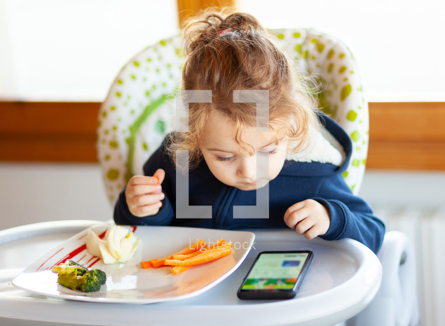 Toddler eats in the high chair while watching movies on the mobile phone