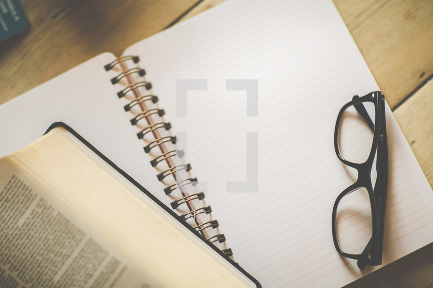 reading glasses on a notebook and an open Bible on a desk 