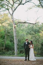 Bride and groom standing on dirt field side of asphalt road with trees in the background.