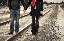 Couple holding hands as they walk along a railroad track.