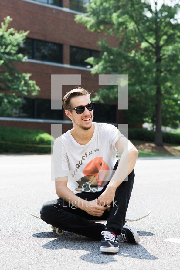Smiling man wearing sunglasses sitting outside on a skateboard.
