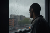 A young man is seen from behind, looking out of a window into the rain