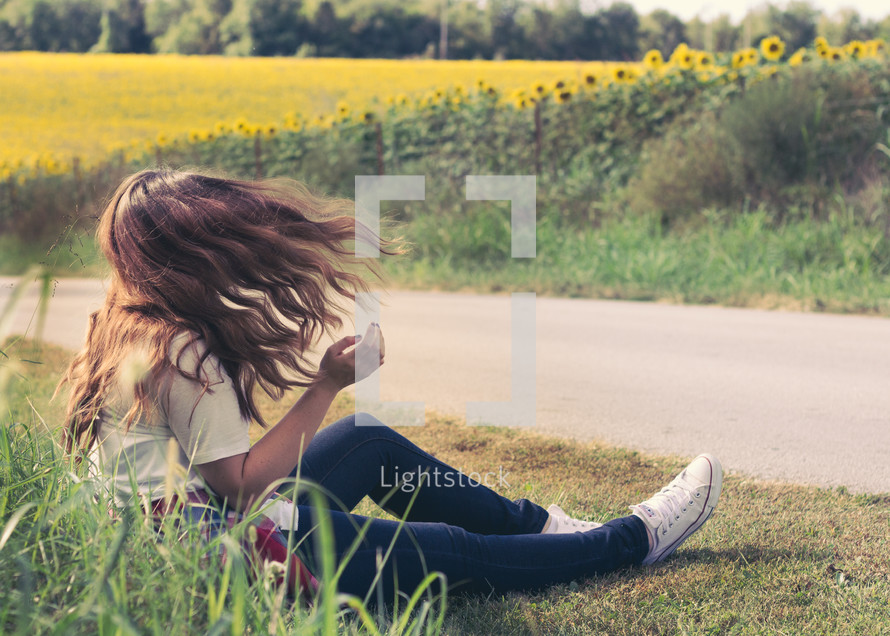 a young woman sitting on the side of a road in front of a field of sunflowers 