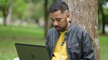 Young student on the grass in the park sits under a tree and cheerfully works with a laptop

