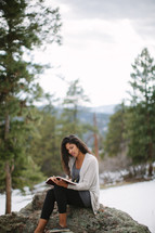 a woman sitting on a rock on a mountaintop reading a Bible 