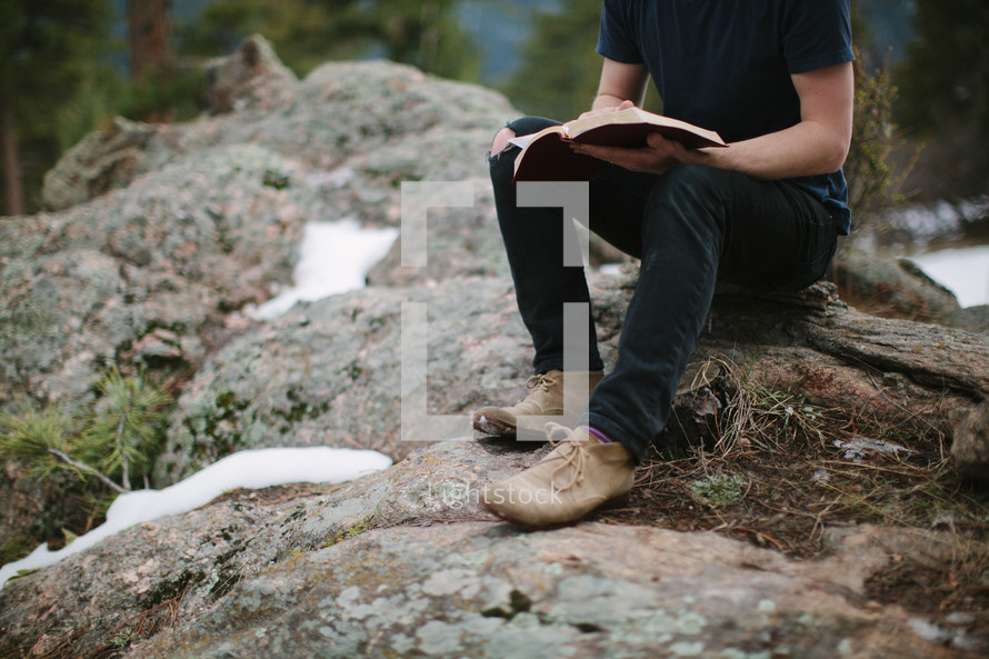 a man sitting on a rock on a mountaintop reading a Bible 