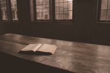 an open Bible on a wooden table in an empty room full of windows 