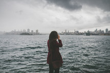 woman standing across the river from a city taking a picture 