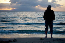 Silhouette of man in jacket with jeans rolled up standing on beach with waves rolling in and cloudy sky at sunset.