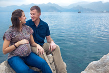 Pregnant woman with man sitting on rock with ocean and moutanins in the background.
