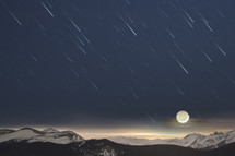 The crescent moon sets over snowy mountains as star trails streak toward the horizon.  