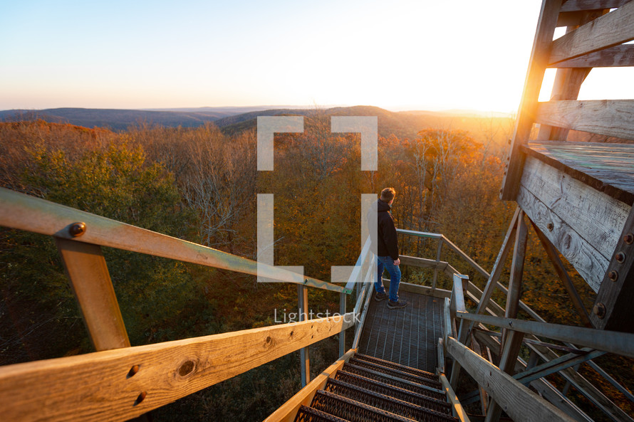Adventurous young man soaks in the sunrise on stairs of wooden watchtower over autumn foliage trees in Bickle Knob West Virginia