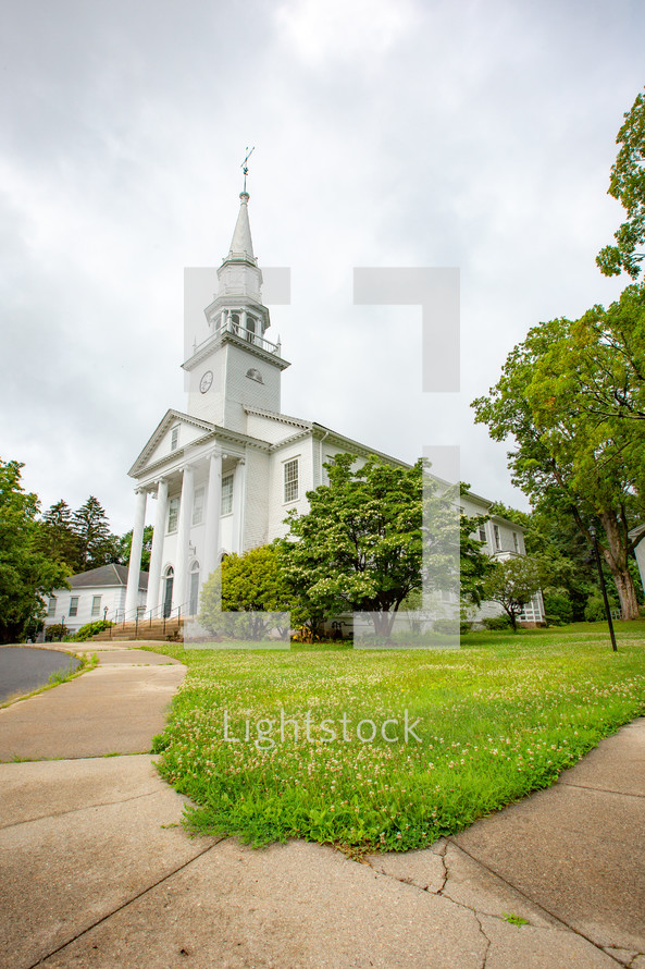 Sidewalk leading to white church building with steeple surrounded by grass and trees