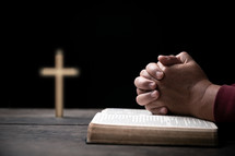 Man's hands folded in prayer over open Bible with cross