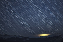 Star trails streak across the night sky toward the snowy mountains on the horizon.  