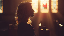 A woman standing in a church alone with light shining through the windows