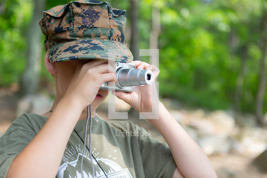 Young boy photographer in army camouflage hat taking picture with point and shoot camera outside 