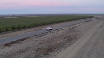 Drone footage of white car on long, empty road near orchards
