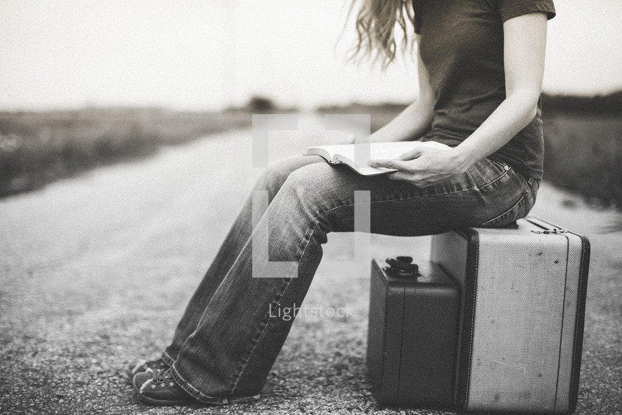 Woman sitting on a suitcase in the middle of the road with an open Bible in her lap.
