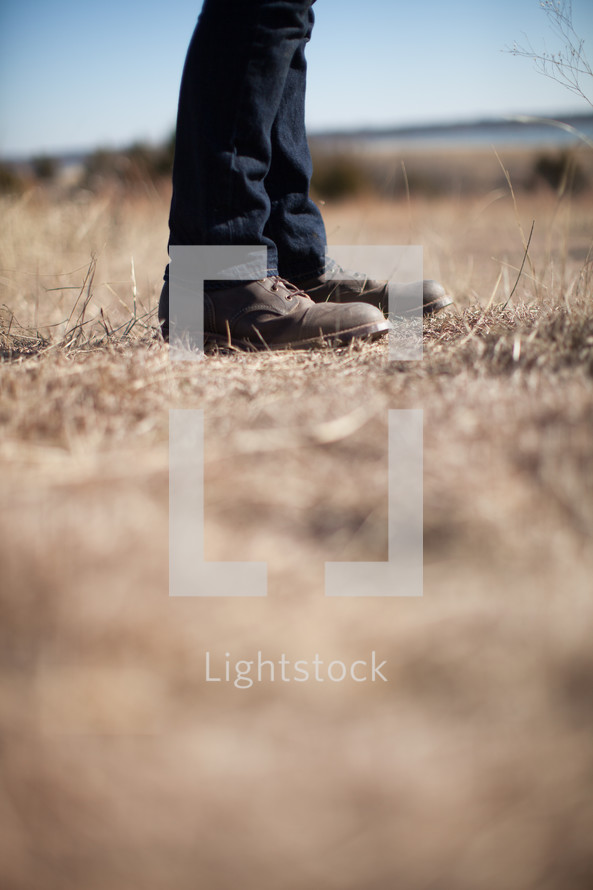 The man's legs in jeans and boots stand in a field of brown grass.
