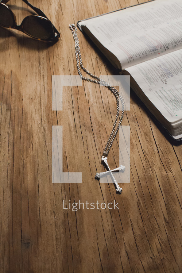A cross on a chain, sunglasses and an open Bible on a wooden tabletop.
