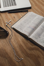 Bible on a wooden table  with a laptop computer, sunglasses, cross pendant, and a pen.