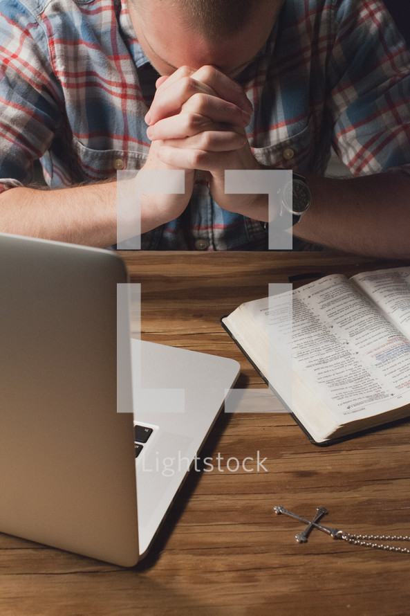 man with his head bowed in prayer by a laptop and Bible 