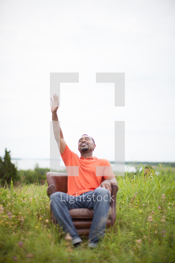 man sitting in a chair in a field with his hand raised to God 