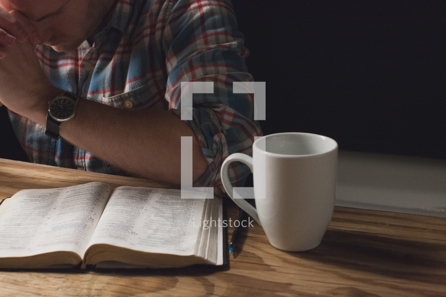 A man praying at a table with an open Bible and coffee cup.