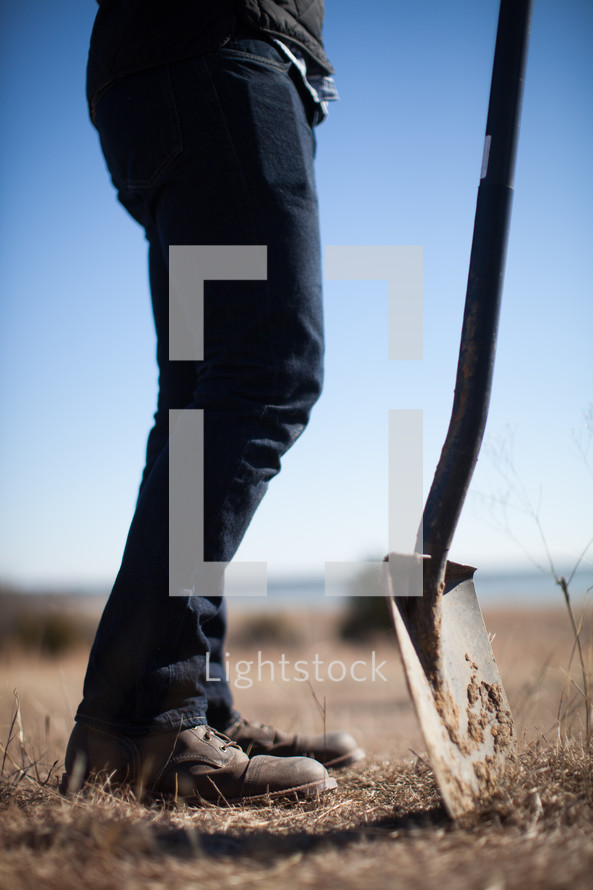 A man stands in a field of brown grass with a shovel.