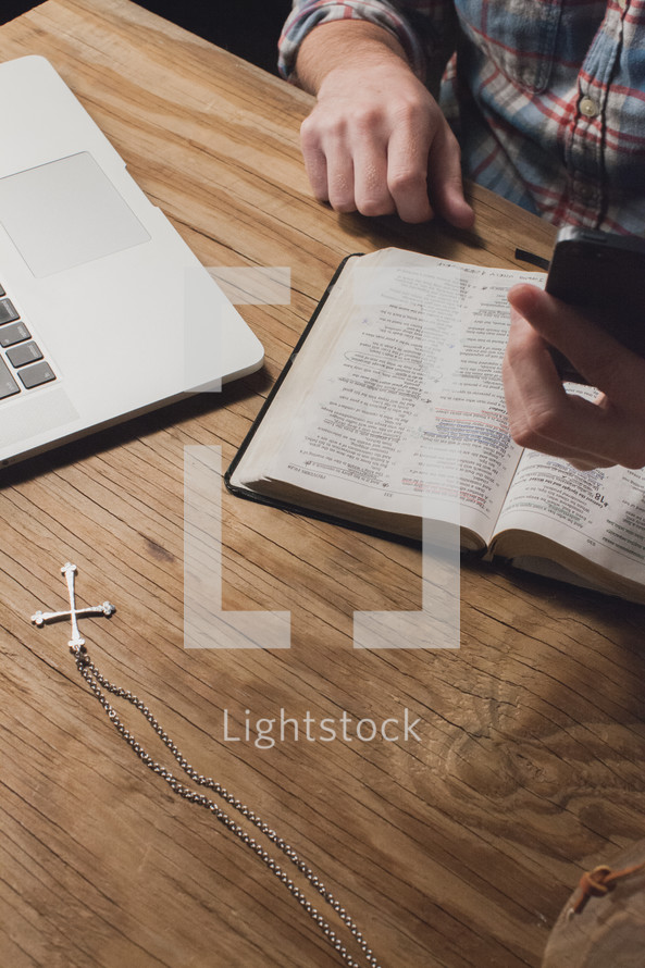 Hand holding a cell phone at a table with a Bible and a laptop computer.
