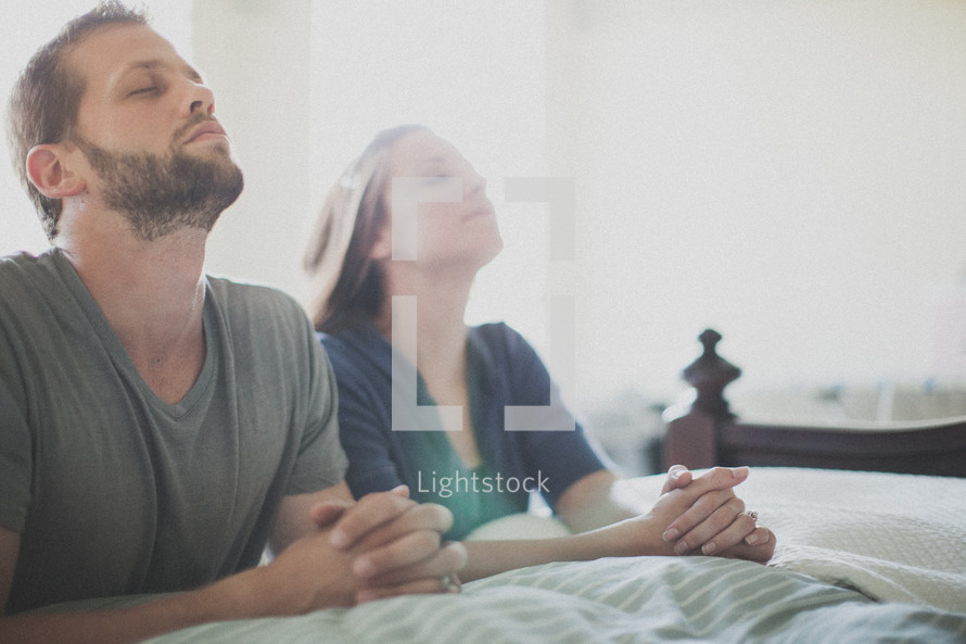 husband and wife praying together at the edge of a bed 