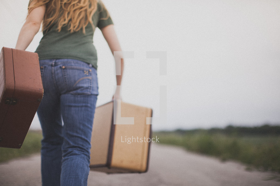 Woman walking down a dirt road carrying suitcases.