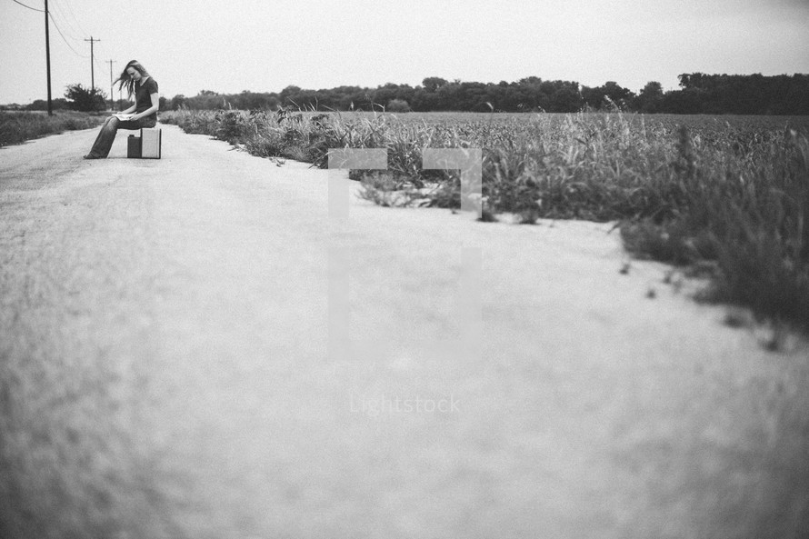 Woman sitting on a suitcase in the middle of a dirt road reading a bible.