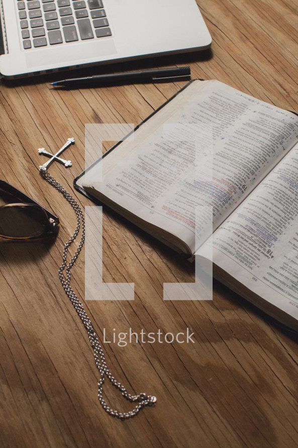 Bible on a wooden table  with a laptop computer, sunglasses, cross pendant, and a pen.